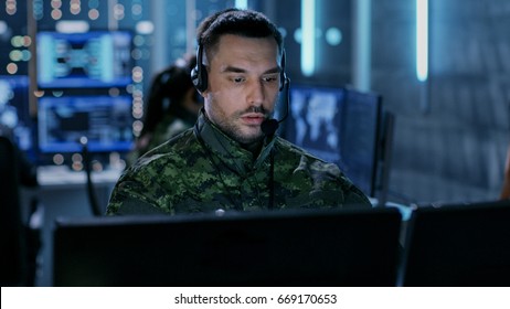 Close-up shot of Military Technical Support Professional Gives Instructions Using Headset. He's in a Monitoring Room with Other Officers and Many Working Displays in Background.