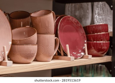 Close-up shot of modern ceramic tableware displayed on a wooden shelf in a shop. Featuring stacked pink mugs with textured patterns, matching bowls, deep burgundy red plates on wooden plate stands, and glass decor in the background.
