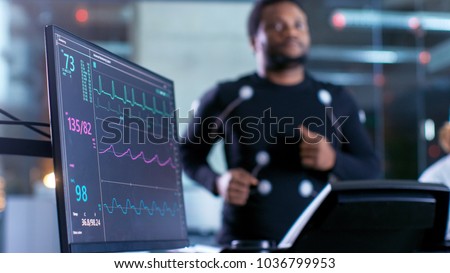 Close-up Shot of a Monitor With EKG Data. Male Athlete Runs on a Treadmill with Electrodes Attached to His Body while Sport Scientist Holds Tablet and Supervises EKG Status in the Background.