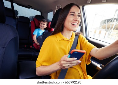 Close-up Shot Of A Mother Driving A Car, Buckled The Seat Belt, Holding Phone. Laughing Boy On Back Seat