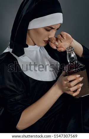 Close-up shot of a nun, sitting on a chair. She's holding bottle of wine in right hand, her left hand is covering the nose. Her eyes are closed. 