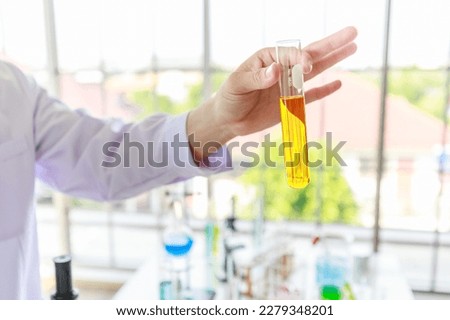 Closeup shot of orange liquid reagent solution in glass test tube holding by unrecognizable unknown scientist hand in white lab coat in front laboratory table with equipment on blurred background.