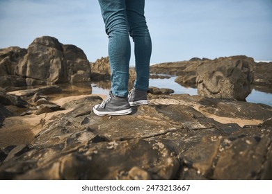 Close-up shot of a person wearing jeans and sneakers standing on a rocky shore. The scene depicts an outdoor adventure by the sea and evokes feelings of exploration and relaxation. - Powered by Shutterstock