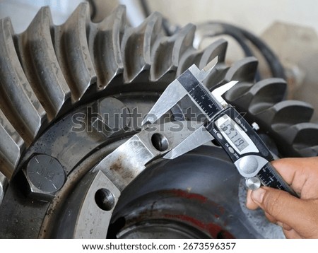 Close-up shot of a person's hand using a digital caliper to measure the dimensions of a large industrial gear. The display shows a precise reading