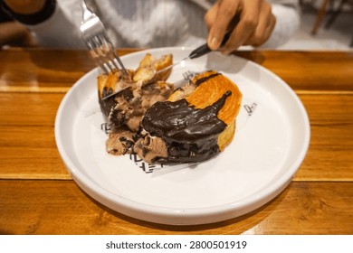 A close-up shot of a person's hands using a fork and knife to cut into a decadent chocolate-covered dessert served on a white plate, set against a warm wooden table.