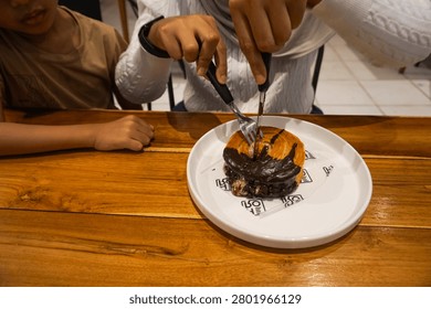 A close-up shot of a person's hands using a knife and fork to cut into a decadent chocolate and orange pancake stack, served on a white plate with unique black lettering, resting on a warm wooden tab.