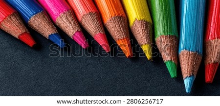 A close-up shot of a row of sharpened colored pencils in various bright hues, arranged diagonally on a dark, textured background.