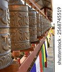 A close-up shot of a row of traditional Buddhist prayer wheels. These metallic cylinders are adorned with intricate carvings and inscriptions, likely containing mantras. The images were taken in Nepal