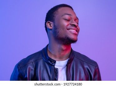 Closeup Shot Of Satisfied Black Guy In Leather Jacket Standing With Closed Eyes Over Purple Studio Background, Pleased Handsome African American Man Posing Under Neon Lighting, Copy Space