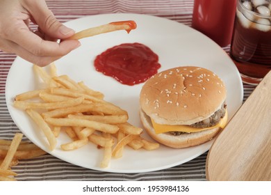 Close-up shot, selective focus: Man's hand was eating French fried, dipping ketchup on a white plate on red tablecloth with hamburgers and cola. Eating junk food or fast food for lunch is unhealthy.