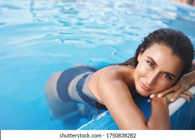 Close-up shot of sexy young tanned woman in bikini, lean swimming pool edge, smiling sensual look camera. Attractive female model in black swimsuit lying in poolside water at hotel, tourism concept.