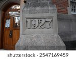 A close-up shot shows a stone column with the number 1927 carved into it. The column is light gray and is situated next to a brown wooden door with a leaded-glass arched window above it.