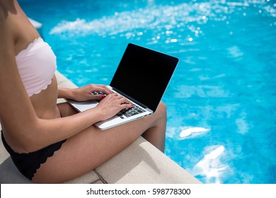 Closeup Shot Of Slim Young Woman In Bikini Sitting With Laptop Near Swimming Pool Outdoors And Dangling Feet In Water. Work At Vacation