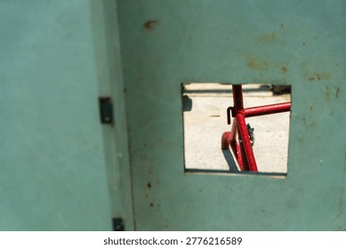 A close-up shot of a small square window cut into a weathered wall, showing part of a red bicycle outside on an outdoor pathway with a rustic urban feel.