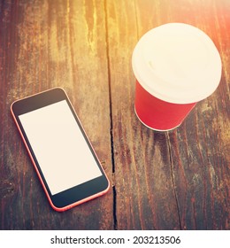 Closeup shot of smart phone white isolated white screen and red cup of takeaway coffee on wooden surface table outdoors. Sunlit. Square image. Instant filter. Contemporary concept. Mock-up. 