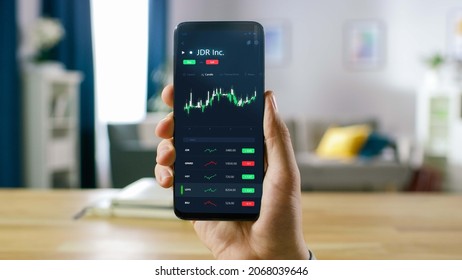 Close-up Shot Of Smartphone. Man Is Using Stock Market App To Check Share Prices. Graph Is Going Up Making A Great Profit. In The Background Cozy Homely Atmosphere.