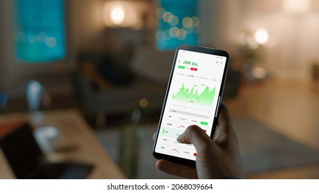 Close-up Shot Of Smartphone. Man Is Using Stock Market App To Check Share Prices. Graph Is Going Up Making A Great Profit. In The Background Cozy Homely Atmosphere, Dusk Time.