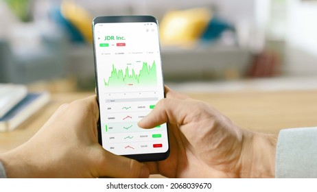 Close-up Shot Of Smartphone. Man Is Using Stock Market App To Check Share Prices. Graph Is Going Up Making A Great Profit. In The Background Cozy Homely Atmosphere.