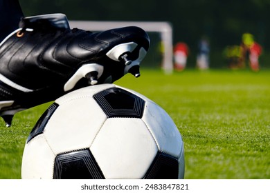 A close-up shot of a soccer player's cleat resting on a black and white soccer ball on a green grass field, with an out-of-focus soccer goal and players in the background. - Powered by Shutterstock