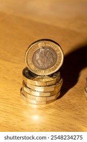 A close-up shot of a stack of British one-pound coins on a light brown wooden surface.  The top coin is a 2016 issue, showing Queen Elizabeth II's profile.