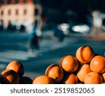 A close-up shot of a stack of oranges in the foreground with a blurred urban street scene in the background, creating a vivid contrast between the vibrant fruits and the soft bokeh of city life.