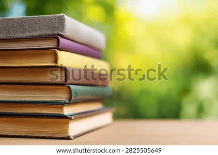 A close-up shot of a stack of weathered, vintage books resting on a wooden surface. The background is a soft, blurred expanse of vibrant green foliage, suggesting an outdoor or natural setting.