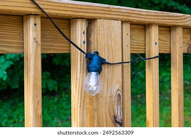 A close-up shot of a string light bulb attached to a wooden fence. The bulb is wet and appears to have been rained on.