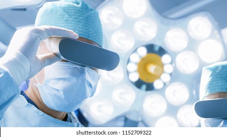 Close-up Shot Of A Surgeon With Augmented Reality Glasses Performing State Of The Art Surgery In High Tech Hospital. Doctors And Assistants Working In Operating Room.