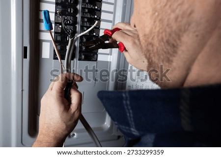 A close-up shot of a technician or handyman using wire strippers to prepare electrical cables for installation in a home circuit breaker box. Concept of home maintenance, electrical safety, and professional trade services.