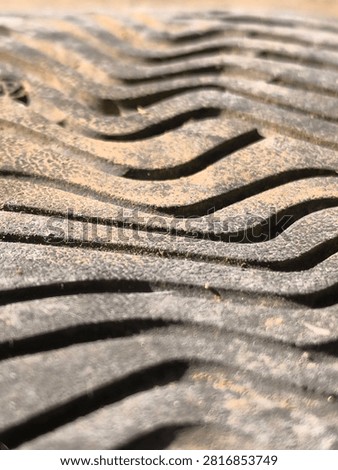 A close-up shot of the textured underside of a white sneaker, featuring a repeating pattern of raised rectangular lugs arranged in rows. The rubber material has a slightly grainy, pebble-like texture, with accumulated dirt settled in the recessed areas between the traction blocks. Sunlight illuminates the scene from an angle, casting distinct shadows that emphasize the three-dimensional depth and height of each lug. The clean white color contrasts with the darker, grayish grime trapped within the crevices, telling a story of use and outdoor activity. The shallow depth of field blurs the distant edges, drawing attention to the precise geometric arrangement of the footwear outsole. This image is well-suited for projects related to athletic footwear, sports equipment, cleanliness versus wear, industrial design, and abstract textural backgrounds. The symmetrical yet organic layout provides a visually pleasing rhythm and pattern suitable for various modern graphic design and commercial application needs.