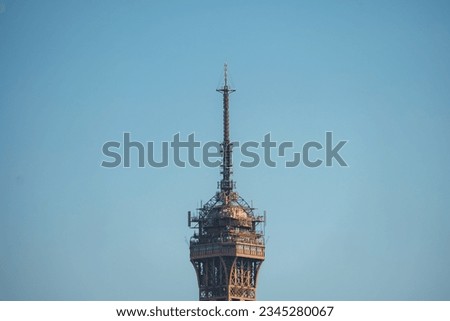 Close-up shot of the top of the Eiffel Tower against a clear blue sky, captured from a sideways angle.