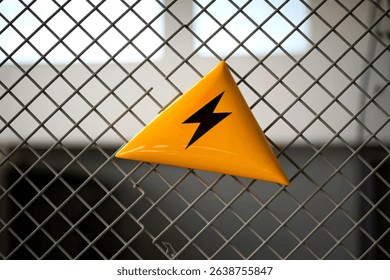 A close-up shot of a triangular yellow warning sign with a black lightning bolt symbol, affixed to a gray metal mesh fence. The sign indicates high voltage danger.