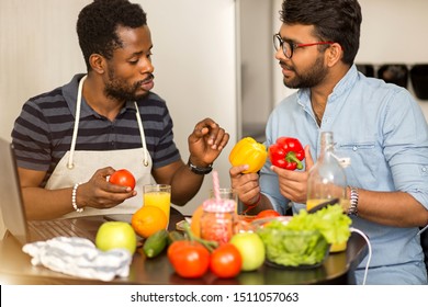 Closeup shot of two multi ethnic bearded friends in casual clothes preparing vegetable salad or smoothie, sitting at table talking using laptop computer in kitchen. Culinary show concept. - Powered by Shutterstock