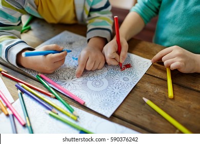 Close-up shot of two pairs of little hands coloring mandala with felt-tip pens, pencils of different colors and illustrations laid on wooden table