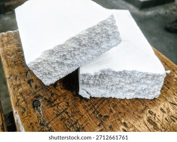 A close-up shot of two rectangular blocks of white styrofoam or expanded polystyrene (EPS) foam, stacked and resting on a rustic, glossy, dark brown wooden surface.