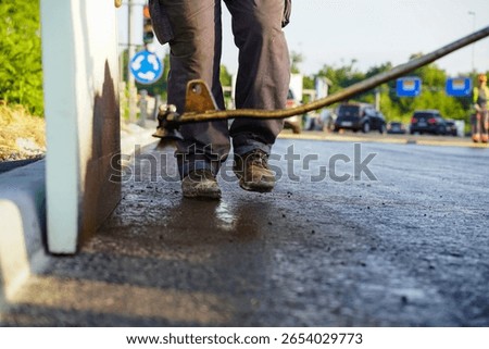 Close-up shot of two workers spraying liquid bitumen onto the edge of freshly laid asphalt next to concrete road curbs at a highway construction site.