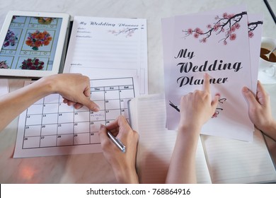 Close-up Shot Of Unrecognizable Couple Wrapped Up In Planning Wedding While Sitting At Table, Man Taking Notes In Calendar