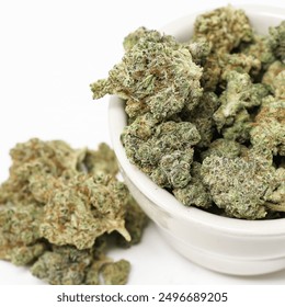 A close-up shot of vibrant cannabis buds spilling out of a small white bowl, showcasing the dense, trichome-covered nugs with their rich green hues and hints of orange and white.