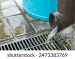 A close-up shot of water pouring from a brown gutter pipe into a metal drain. The water is flowing freely and the drain is surrounded by concrete pavers.