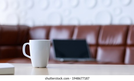 Closeup Shot Of White Coffee Mug Placed On Left Side Of Wooden Table In Front Of Brown Leather Cozy Sofa, Black Laptop Computer Monitor And White Circle Shape Surface Wall In Blurred Background.