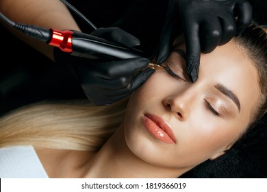 Closeup shot of a woman in black gloves making permanent eyelid makeup to a young woman client in beautician salon. Top view