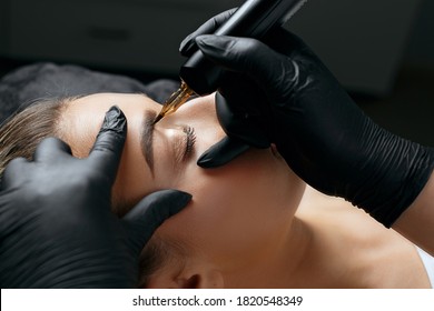 Closeup shot of a woman in black gloves making permanent brow makeup to a young woman in beauty salon