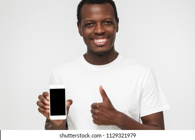 Closeup Shot Of Young African American Man Isolated On White Background Wearing White T Shirt, Showing Blank Phone Screen And Thumb Up Willing To Recommend Product Or Service