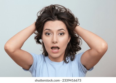 a close-up shot of a young, beautiful, surprised, puzzled woman with brown eyes with curly hair in a blue T-shirt, who looking into the camera with her mouth open and holding her head with her hands - Shutterstock ID 2339416673