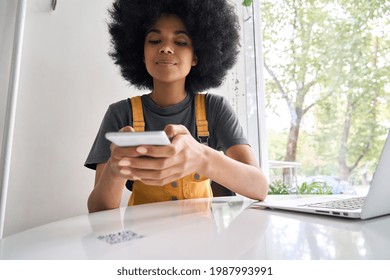 Closeup Shot Of Young Black Gen Z African American Girl With Afro Hair Holding Cell Smart Phone Scanning Qr Code To Read Menu Or Make Payment Online In Cafe Sitting At Table Indoors.