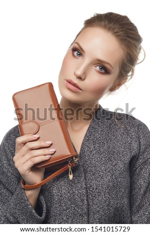 Close-up shot of a young brown-haired European lady in a gray mottled jacket with a textured brown leather wallet with a buckle opening closure and a wristlet strap handle in her hand