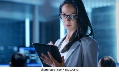 Close-up Shot Of Young Female Government Employee Wearing Glasses Uses Tablet In System Control Center. In The Background Her Coworkers Are At Their Workspaces With Many Displays Showing Valuable Data