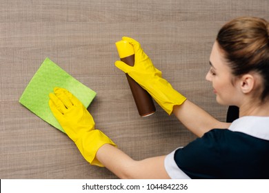 Close-up Shot Of Young Maid In Uniform Wiping Wood Wall With Rag And Aerosol Furniture Cleaner