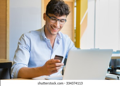 Closeup shot of a young man typing a text on mobile phone. Guy holding a modern smartphone and writing a phone message. Smiling young businessman looking at cellphone with laptop on table.
