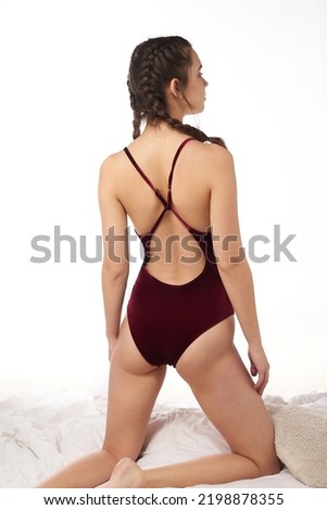 Close-up shot of a young woman in a burgundy one piece velvet swimsuit. The woman with braids in an open back swimsuit is posing on a bed on a white background. Back view.                           